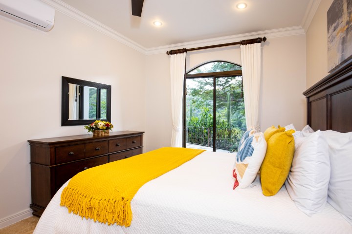 Bedroom with a bed, yellow throw, pillows, dresser, mirror, and window view of greenery.