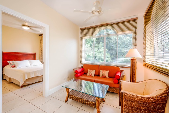 Living room with a sofa, glass table, and view into a bedroom with a bed.