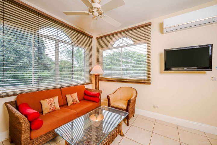 Cozy living room with a wicker sofa, armchair, glass table, and wall-mounted TV. Large windows with blinds.