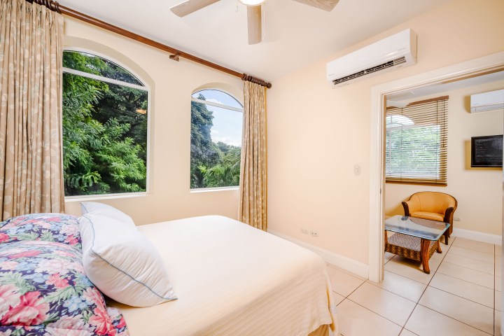 Bedroom with large windows, a double bed, beige curtains, and an adjacent room with chair and table.