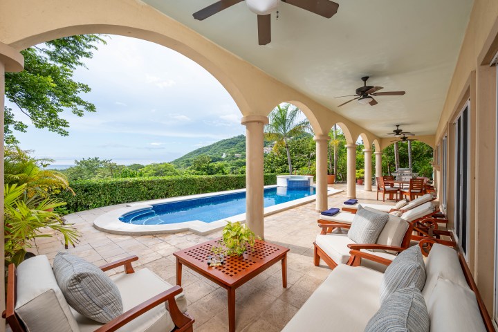 Covered patio with sofas, ceiling fans, arches, and pool overlooking lush landscape and ocean in the distance.