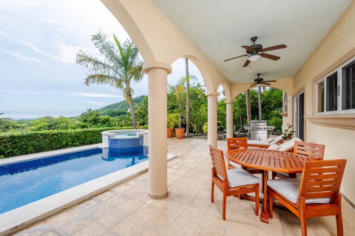 Outdoor patio with pool, round table, chairs, arches, and palm trees, overlooking a green landscape.