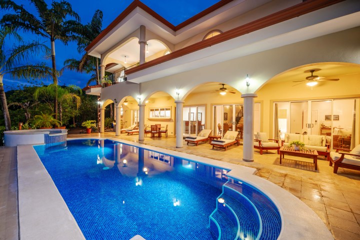 Luxury villa with arched patio, pool, seating, and palm trees at dusk.