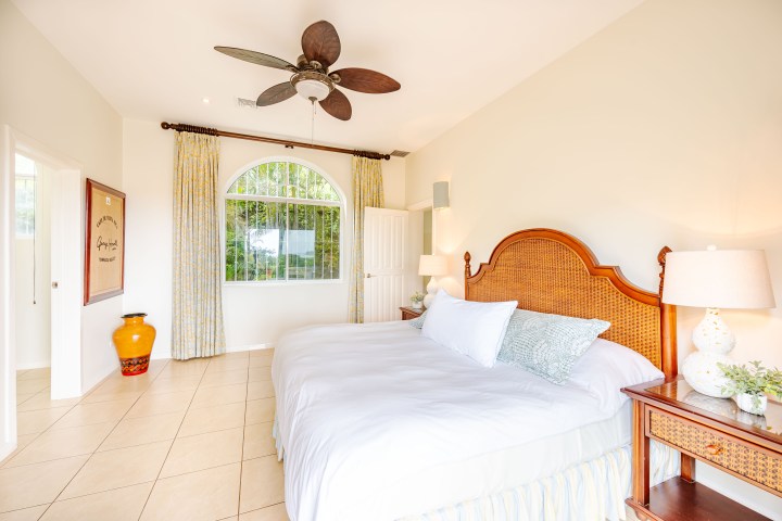 Bright bedroom with king-size bed, wicker headboard, ceiling fan, window, curtains, lamps, and side table.