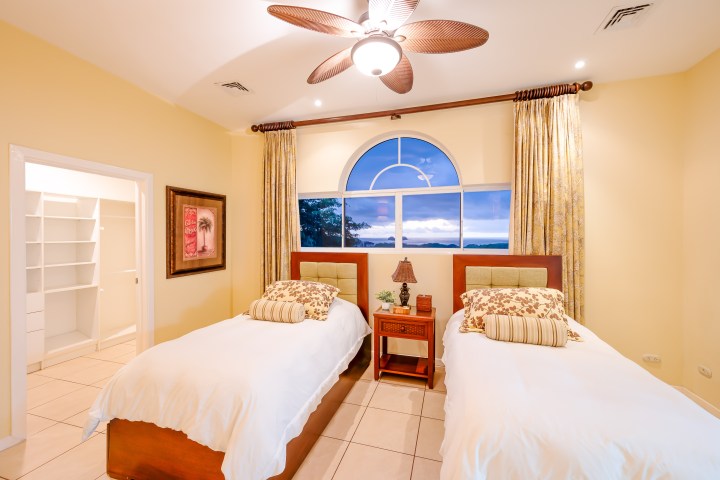 Bright bedroom with two twin beds, ceiling fan, large window, and a view outside.