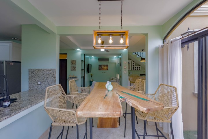 Modern dining room with wooden table, wicker chairs, pendant lights, and green walls.