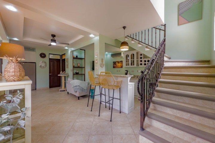 Modern living room with beige tiles, staircase, kitchen island, wicker stools, and wall art.
