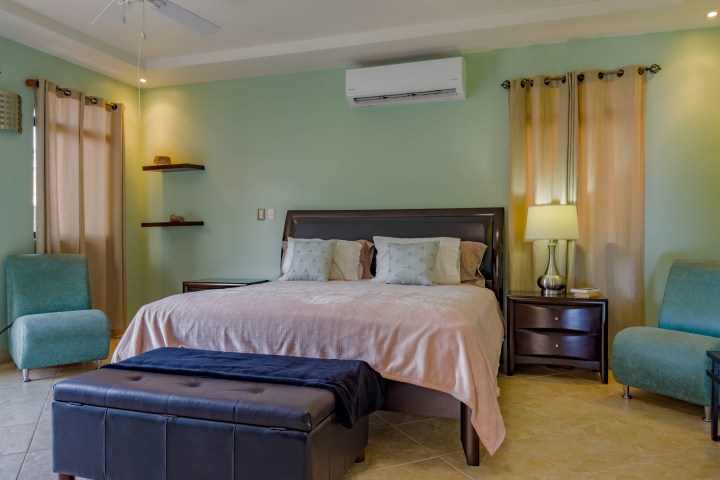 Modern bedroom with a bed, two teal chairs, lamps, and air conditioning on green walls.