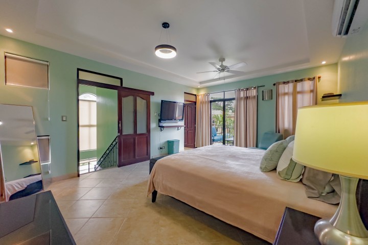 Spacious bedroom with bed, mirror, TV, open door to balcony, ceiling fan, and curtains.