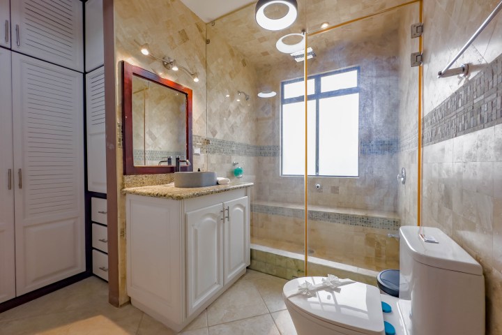 Modern bathroom with beige tiles, glass shower, white cabinets, and a round mirror above the sink.