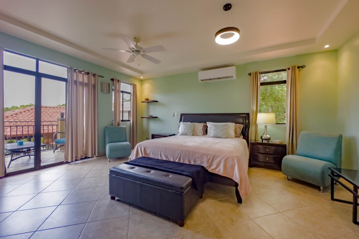 Spacious bedroom with a bed, chairs, a ceiling fan, lamp, and balcony view.