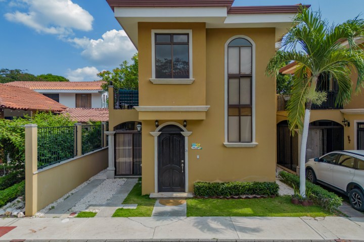 Two-story beige house with dark door and windows, tropical tree on the right, and parked white car.