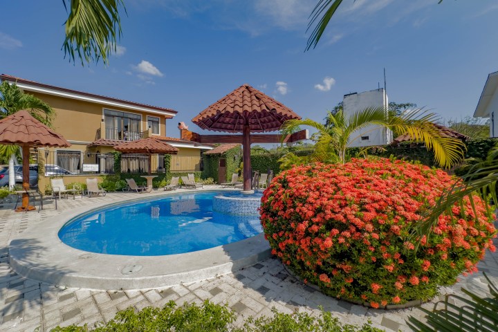 Outdoor pool with red umbrellas, surrounded by flowers and chairs, near a house.