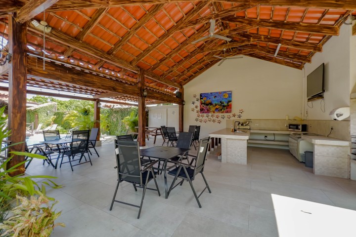 Open-air pavilion with tables, chairs, and a tiled roof beside a kitchen area and greenery.