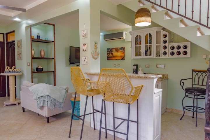 Cozy living area with a bar, wicker chairs, and staircase in a pastel-colored room.