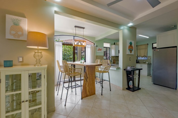Modern kitchen with bar stools at a wooden table, pendant lights, and a view of the garden through large windows.