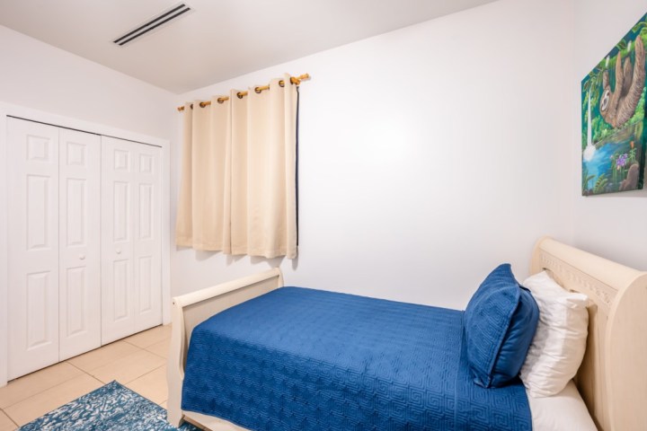 Simple bedroom with a single bed, blue bedding, cream curtains, and a sloth painting on the wall.