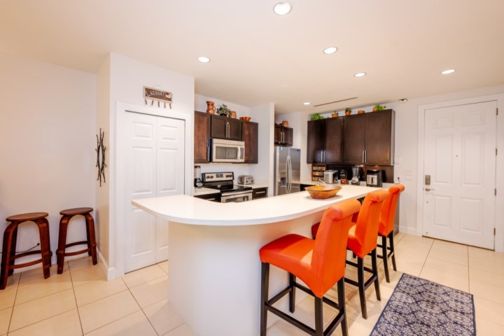 Modern kitchen with white counters, orange stools, stainless steel appliances, and dark cabinets.