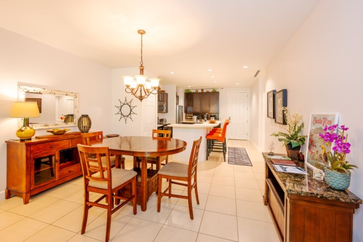Open dining area with wooden table, chairs, chandelier, and kitchen in background.