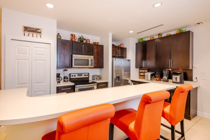 Modern kitchen with dark cabinets, stainless appliances, and three orange bar stools at a white counter.
