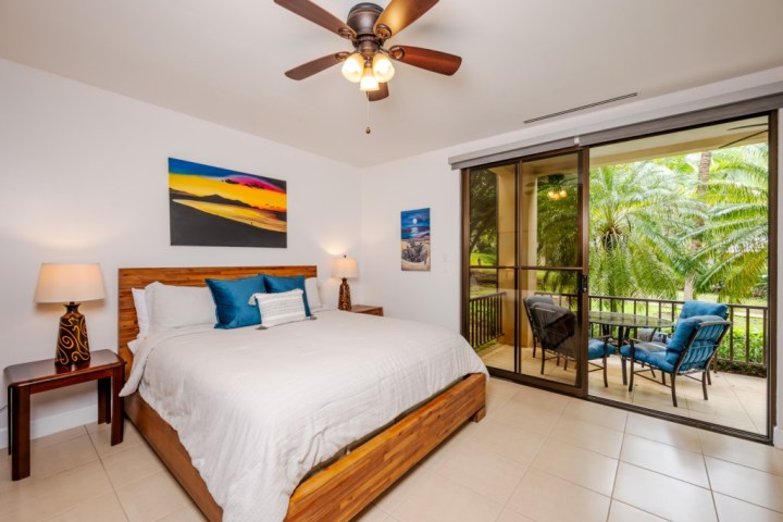 Bedroom with large bed, ceiling fan, art on walls, and sliding doors to balcony with chairs.