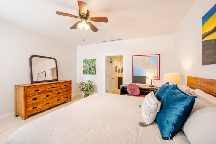 Bedroom with wooden dresser, mirror, ceiling fan, desk with computer, and a bed with blue pillows.
