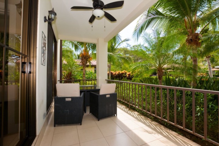 Covered patio with wicker chairs and a table, overlooking lush garden with palm trees.
