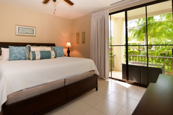 Bedroom with king bed, blue pillows, lamp, sliding glass doors, and view of tropical plants outside.