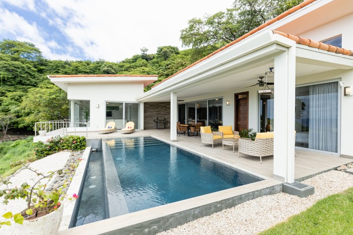 Modern house with a pool, patio furniture, and surrounding greenery under a blue sky.