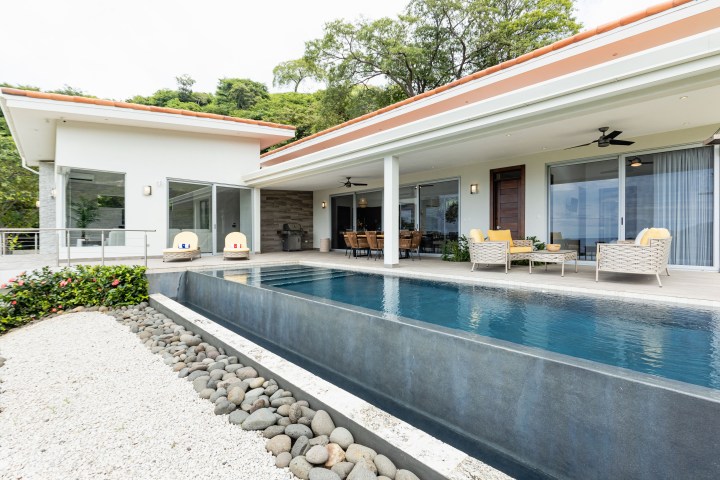 Modern house with large patio, pool, and outdoor furniture surrounded by greenery.