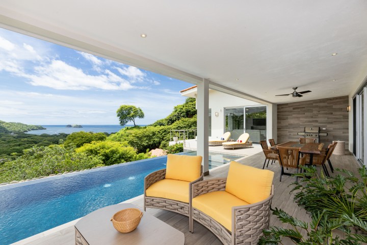 Luxury terrace with pool, ocean view, yellow seating, dining area, and greenery under a clear blue sky.