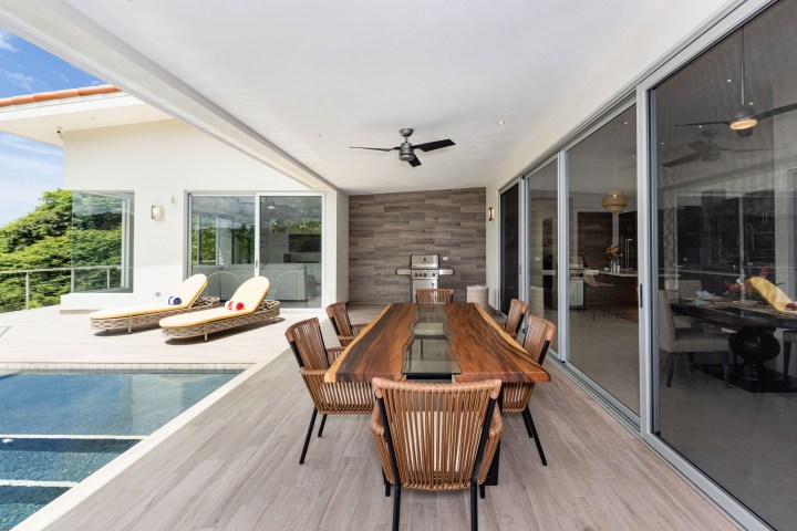 Outdoor patio with a wooden dining table, chairs, and lounge chairs by a pool.