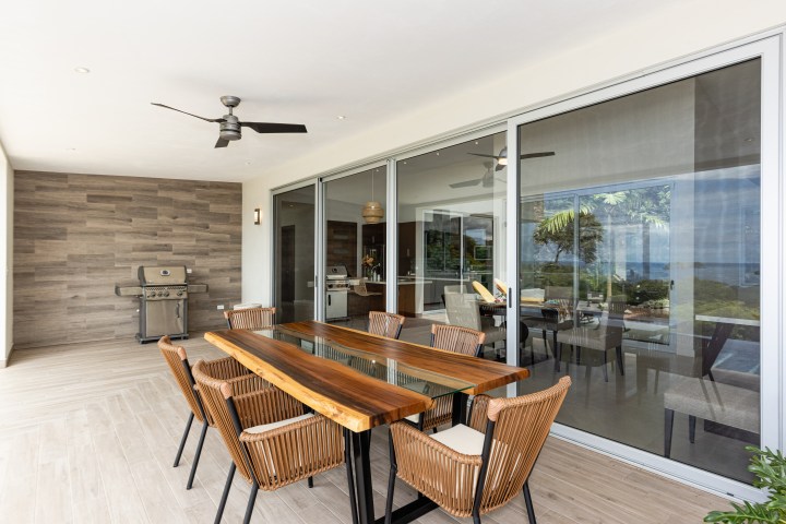 Outdoor dining area with wooden table, wicker chairs, ceiling fan, and a grill on a patio.