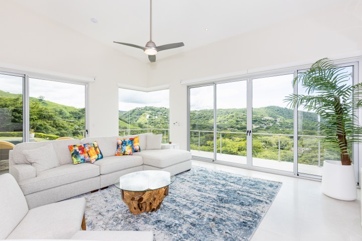 Bright living room with gray sofa, colorful pillows, and large windows showing green mountain views.