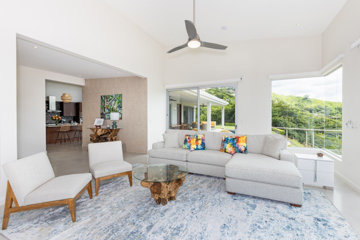 Modern living room with gray sofa, colorful pillows, glass table, and large windows with greenery outside.