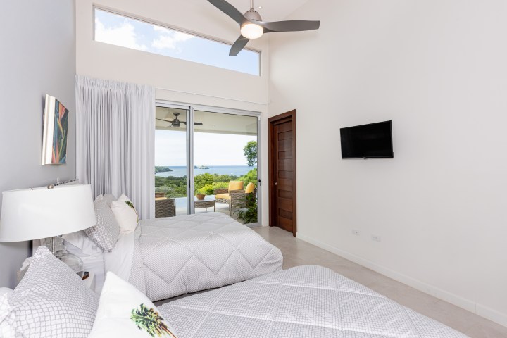 Bedroom with two beds, wall-mounted TV, and glass doors opening to a balcony with ocean view.
