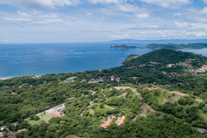 Aerial view of lush green landscape, coastal shoreline, and distant hills under a cloudy blue sky.