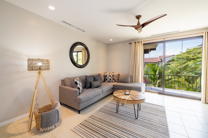 Modern living room with a gray sofa, striped rug, round table, and large window viewing greenery.