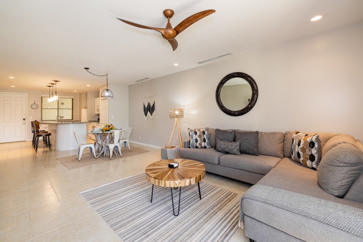 Open living room with gray sofa, round table, fan, and kitchen at the back.
