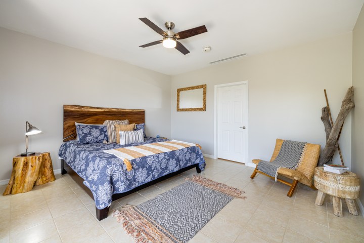 Cozy bedroom with a bed, wooden table, ceiling fan, chair, and decorative items on a tiled floor.