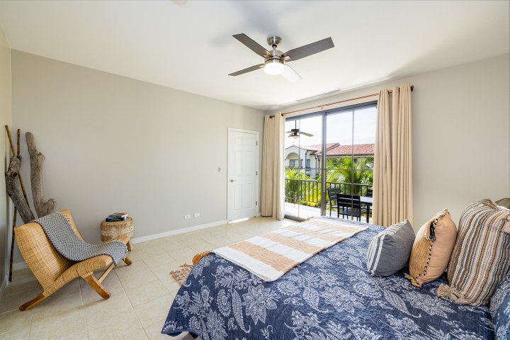 Bedroom with large window, ceiling fan, and striped bedspread. Chair and table on left, view of balcony with palm trees.