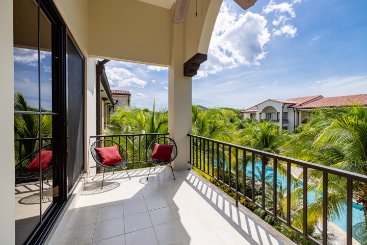 Balcony with two red-cushioned chairs, overlooking tropical trees and buildings.