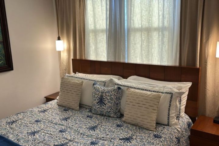 Cozy bedroom with blue floral bedding, wooden headboard, two pendant lights, and a parrot painting on the wall.
