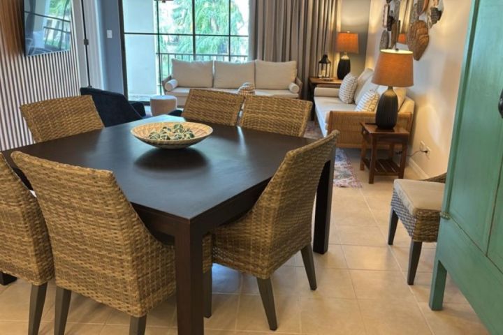 Dining table with wicker chairs near a cozy living area with sofas and lamps.