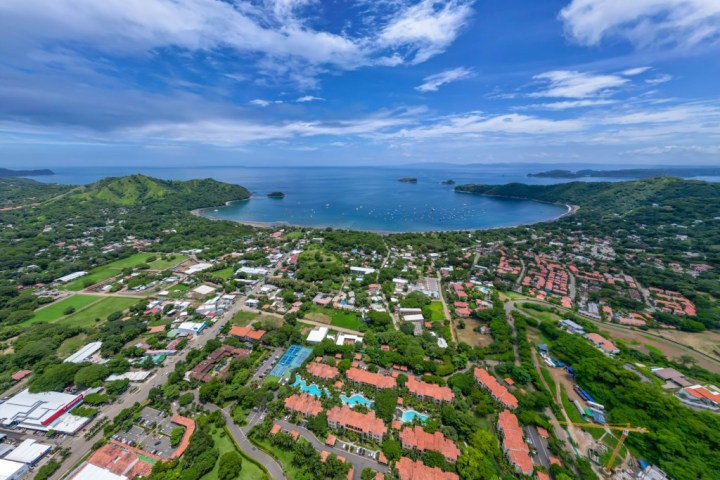 Aerial view of coastline with bay, green hills, and scattered buildings under a blue sky.