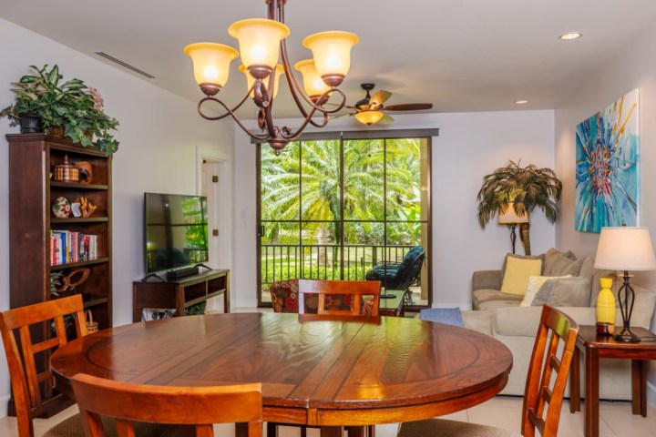 Cozy living area with wooden dining table, sofa, TV, bookshelf, and large window overlooking greenery.