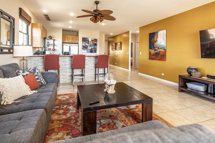 Living room with gray sofa, colorful rug, and open kitchen with bar stools and yellow walls.