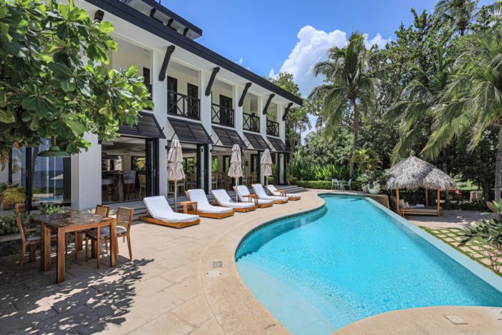 Modern villa with a pool, sun loungers, palm trees, and outdoor dining area on a sunny day.