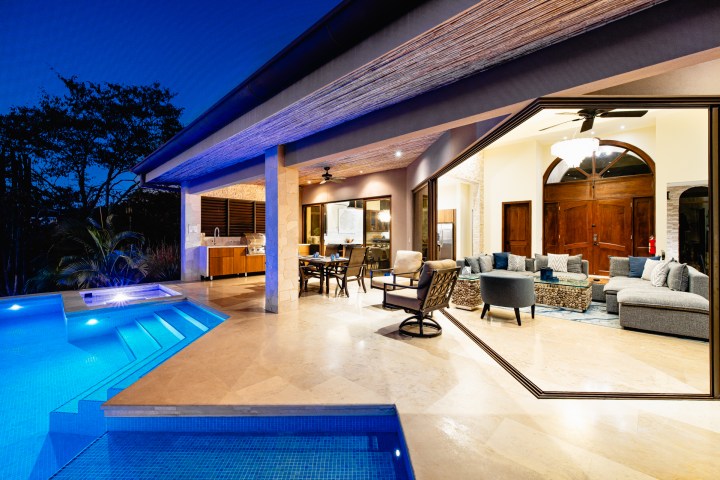Modern open-concept home with lit pool, outdoor kitchen, and stylish indoor living area at night.