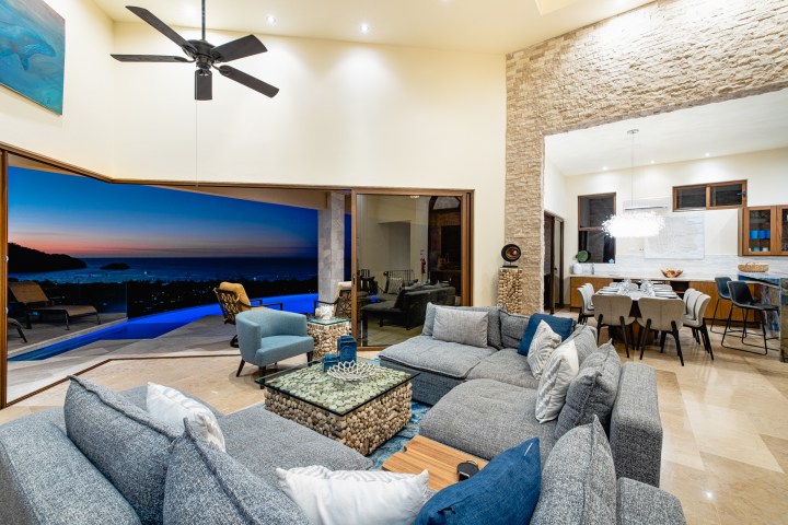Modern living room with ocean view at sunset, large sofas, dining table, and ceiling fan.